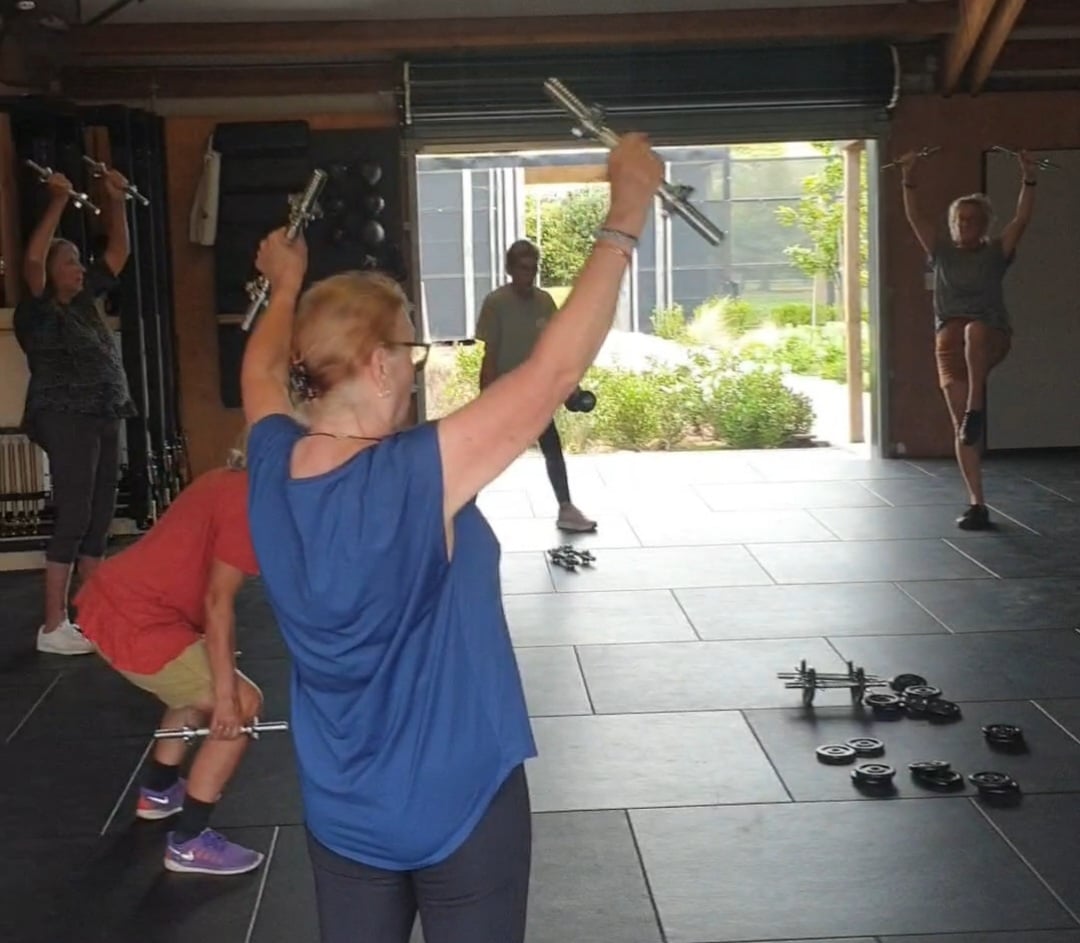 An over-60s class working with weighted bars overhead, coach watching from the open roller door